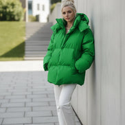 A woman wearing an oversized bright green puffer jacket with a high collar and front zipper, paired with white pants, standing against a textured brown wall.
