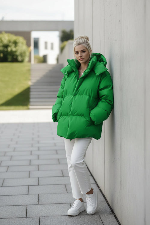 A woman wearing an oversized bright green puffer jacket with a high collar and front zipper, paired with white pants, standing against a textured brown wall.