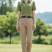 A woman wearing a short-sleeve olive green polo shirt with white polka dots, paired with beige trousers and a black belt with a silver buckle.