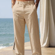 A man wearing light beige linen trousers stands barefoot on a patio with the ocean and a white draped curtain in the background.