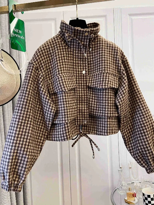 Brown and cream houndstooth utility jacket on a hanger, showing a raised drawcord collar, chest flap pockets, front snap buttons, elasticated cuffs and a tied drawcord hem