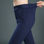 A close-up of a person wearing navy blue tailored trousers, holding the waistband to show the fit and stretch. The background is a solid dark gray studio backdrop.