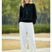 A woman wearing a black long-sleeve knit sweater with a crew neck, paired with white pants and a silver pendant necklace.