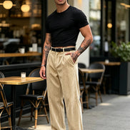 A man stands on a city sidewalk in front of a cafe, wearing beige corduroy trousers, a black t-shirt, a black belt, and black-and-white loafers.