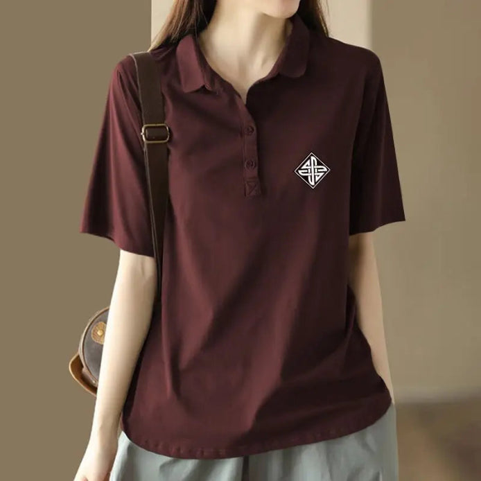 A woman wearing a short-sleeve polo shirt in deep burgundy with a small white geometric logo on the left chest, paired with light gray pants and a brown shoulder bag.