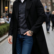 A man wearing a long black double-breasted overcoat over a dark gray button-down shirt and blue jeans, walking on a city sidewalk.