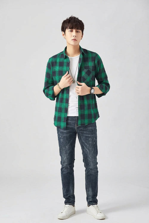 A man modeling a green and black plaid button-up shirt over a white t-shirt and dark blue jeans, set against a plain light gray studio background.