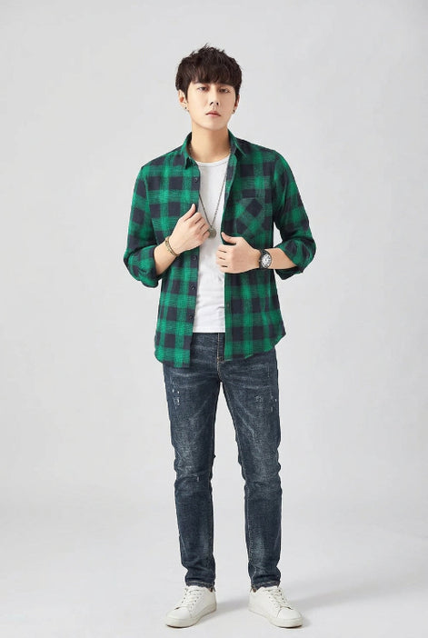 A man modeling a green and black plaid button-up shirt over a white t-shirt and dark blue jeans, set against a plain light gray studio background.
