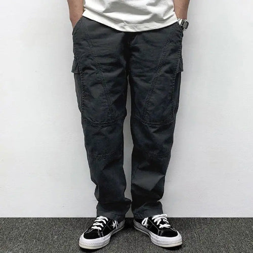 Front view of a model wearing dark gray loose-fit cargo pants with thigh pockets, curved seam detailing, and stitched knee panels, paired with black sneakers.
