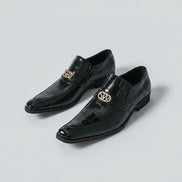 Black dress shoes with gold logo worn by a person on a white background