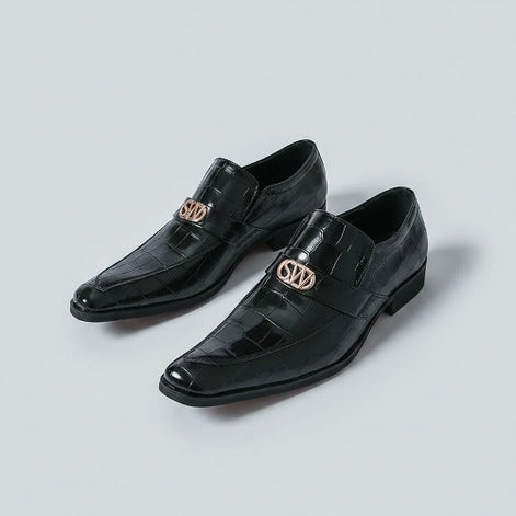 Black dress shoes with gold logo worn by a person on a white background