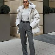 A woman wearing a light gray oversized puffer jacket, gray knit beanie, gray sweatshirt, and gray trousers, standing outdoors 