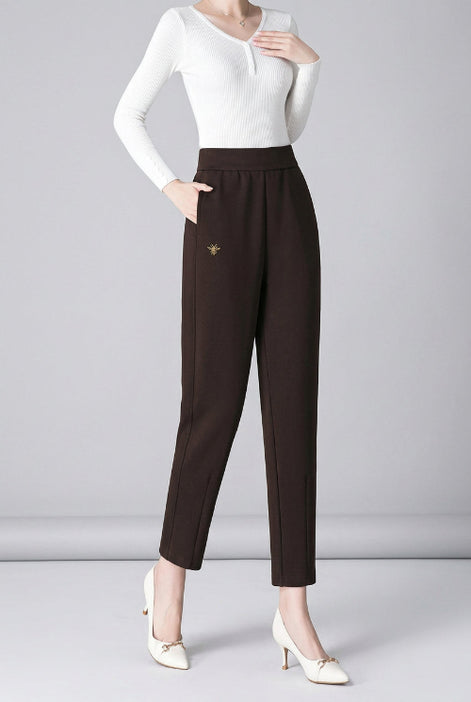 A woman models a pair of slim-fit, high-waisted brown trousers with a small gold embroidered bee detail on the left hip. 