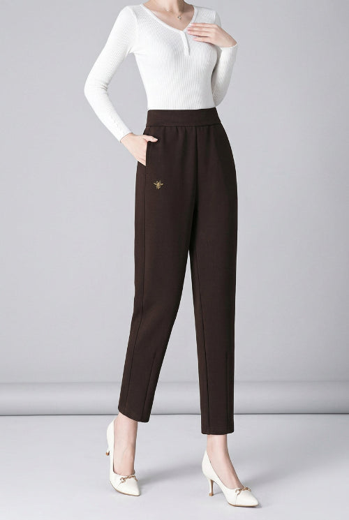 A woman models a pair of slim-fit, high-waisted brown trousers with a small gold embroidered bee detail on the left hip. 