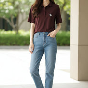 A woman wearing a short-sleeve polo shirt in deep burgundy with a small white geometric logo on the left chest, paired with a dark brown shoulder bag strap.