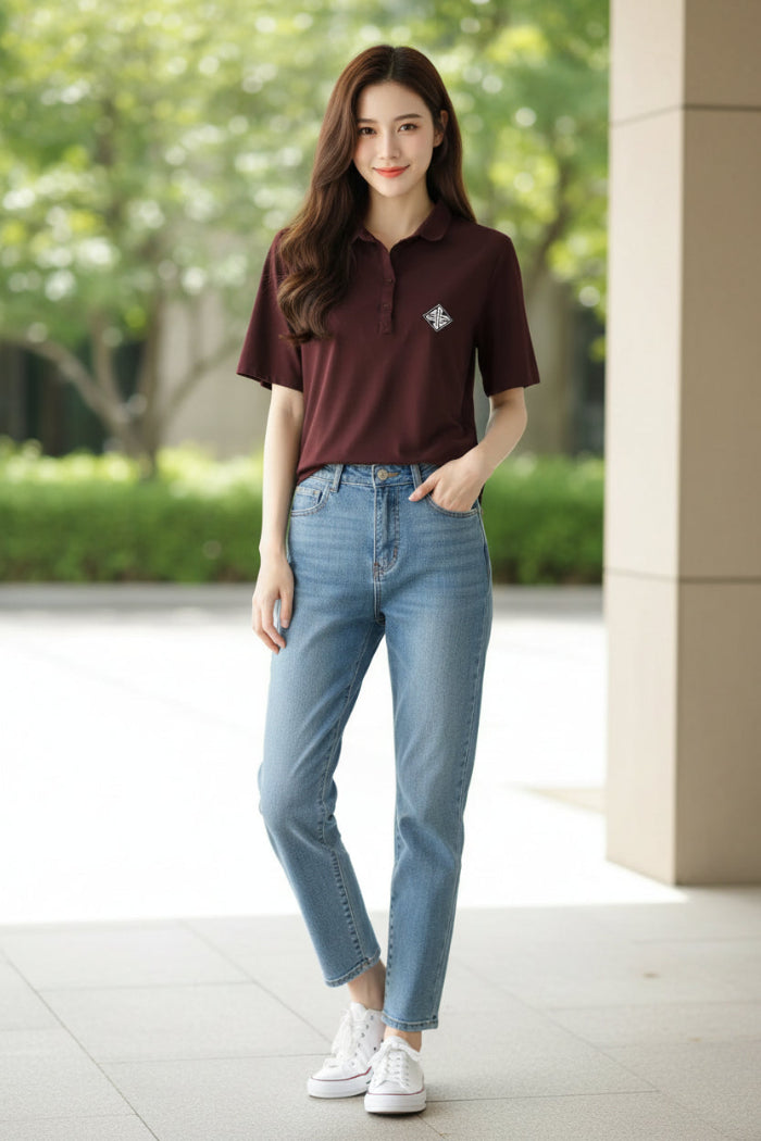 A woman wearing a short-sleeve polo shirt in deep burgundy with a small white geometric logo on the left chest, paired with a dark brown shoulder bag strap.