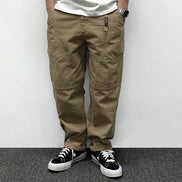 Front view of a model wearing khaki loose-fit cargo pants with thigh pockets, curved seam detailing, and stitched knee panels, paired with black sneakers.