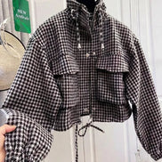 Black and white houndstooth utility jacket on a hanger, showing a raised drawcord collar, chest flap pockets, front zipper and snaps, elasticated cuffs and a drawcord hem