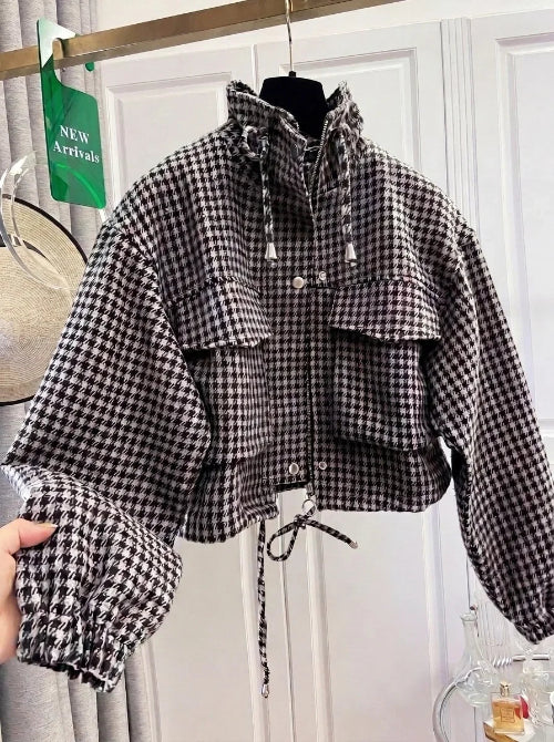 Black and white houndstooth utility jacket on a hanger, showing a raised drawcord collar, chest flap pockets, front zipper and snaps, elasticated cuffs and a drawcord hem