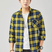 A man wearing a yellow and blue plaid button-up shirt over a white t-shirt and black jeans, standing against a light gray studio background.