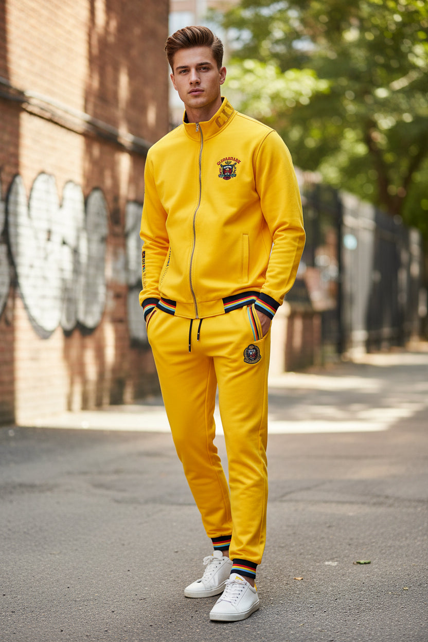 A bright yellow two-piece tracksuit set consisting of a zip-up jacket and matching pants, featuring multicolor striped ribbed cuffs and hem, a central crest logo on the chest, and side zipper pockets.
