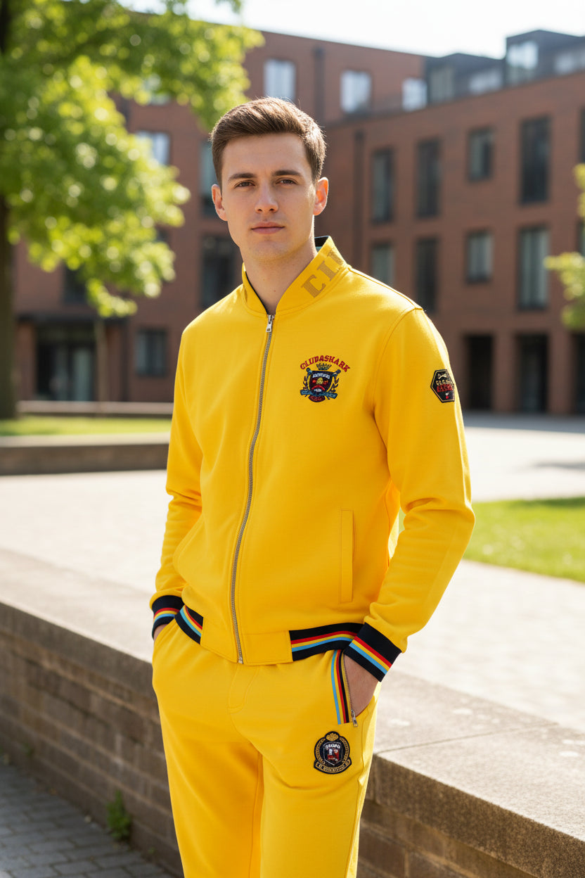 A bright yellow two-piece tracksuit set consisting of a zip-up jacket and matching pants, featuring multicolor striped ribbed cuffs and hem, a central crest logo on the chest, and side zipper pockets.