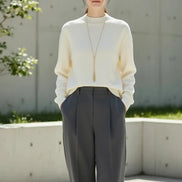 A woman wearing a cream-colored ribbed knit mock-neck sweater with a long gold pendant necklace, paired with a black skirt. 