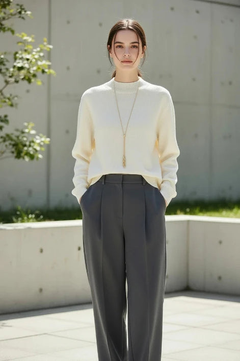 A woman wearing a cream-colored ribbed knit mock-neck sweater with a long gold pendant necklace, paired with a black skirt. 