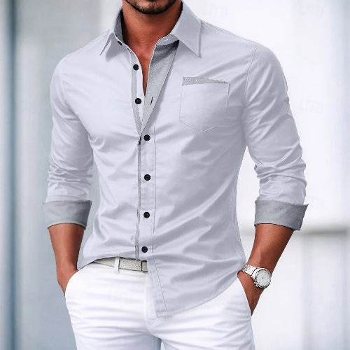 Man wearing a light gray dress shirt with black buttons and white pants against a blurred background