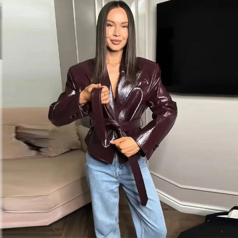 Woman tying the waist belt of a glossy dark brown jacket with wide lapels and buttoned cuffs, styled with light blue jeans