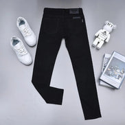 Black slim-fit jeans laid flat on a light gray surface, styled with white sneakers, a small bear figurine, and a stack of magazines.