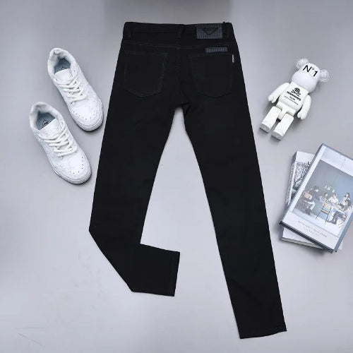 Black slim-fit jeans laid flat on a light gray surface, styled with white sneakers, a small bear figurine, and a stack of magazines.