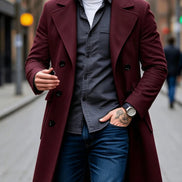 A man wearing a long maroon double-breasted overcoat over a dark gray button-up shirt, white t-shirt, and blue jeans, walking on a city street.