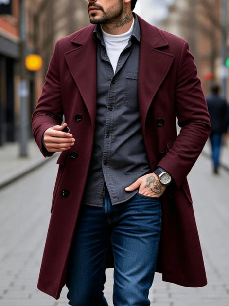 A man wearing a long maroon double-breasted overcoat over a dark gray button-up shirt, white t-shirt, and blue jeans, walking on a city street.