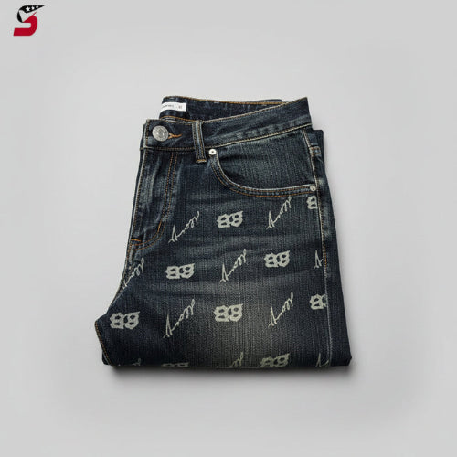 A hand holding a pair of folded dark blue denim jeans with a repeating light beige 'BB' monogram pattern, 