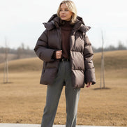 Woman wearing a brown puffer jacket in a room with a white wall and light-colored furniture.