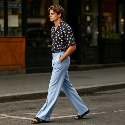 A man walking on a city street wearing a black short-sleeve button-up shirt with white polka dots, light blue wide-leg trousers, and black slide sandals.