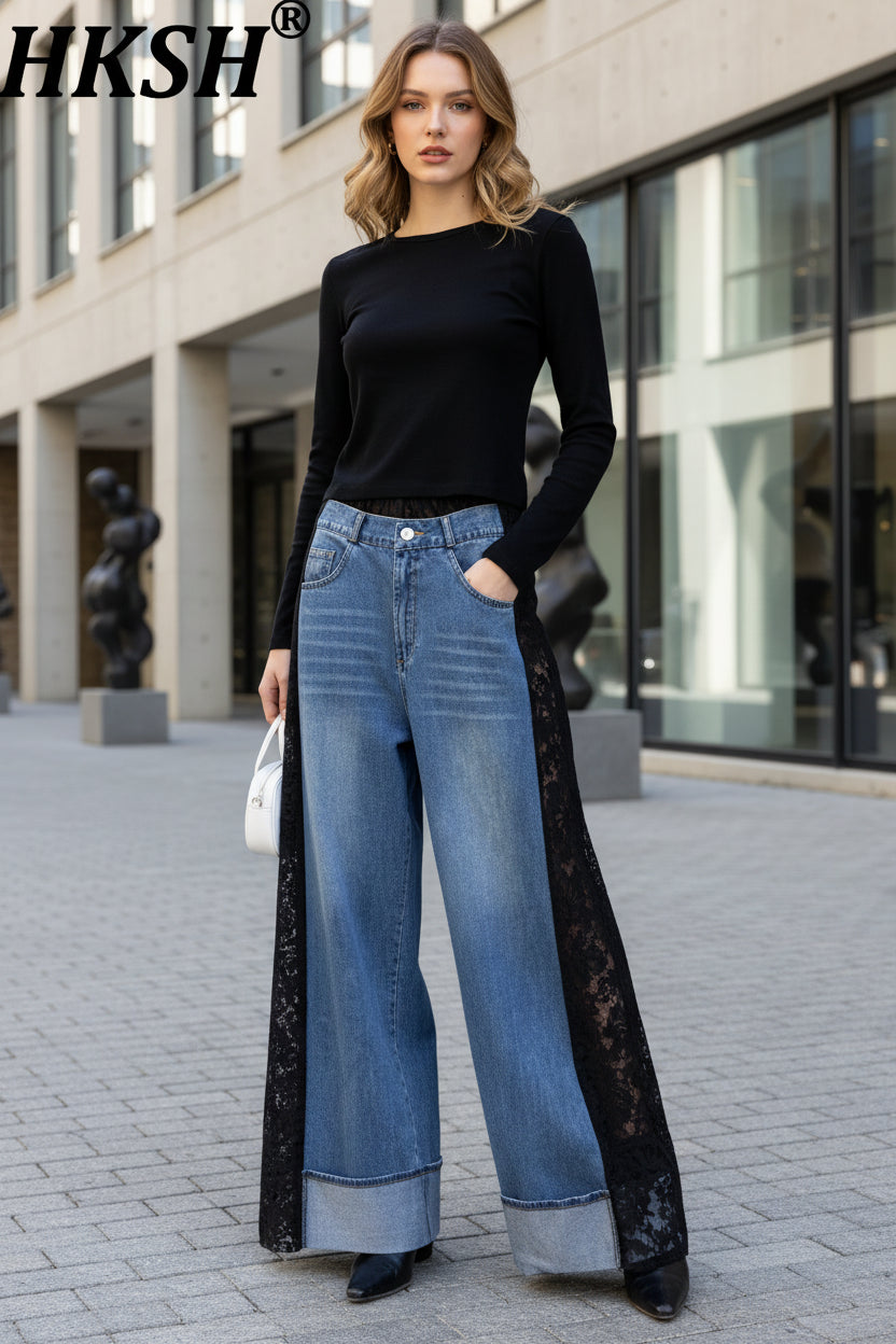 A person wearing HKSH wide-leg blue denim jeans with black lace side panels, styled with a black long-sleeve top and black shoes, standing against a light textured wall.