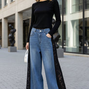 A person wearing HKSH wide-leg blue denim jeans with black lace side panels, styled with a black long-sleeve top and black shoes, standing against a light textured wall.