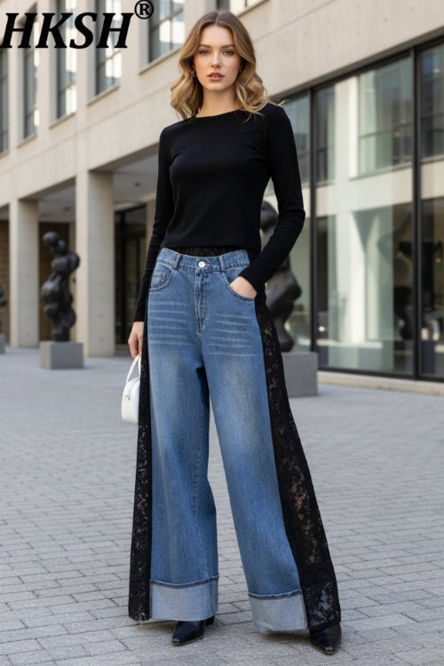 A person wearing HKSH wide-leg blue denim jeans with black lace side panels, styled with a black long-sleeve top and black shoes, standing against a light textured wall.