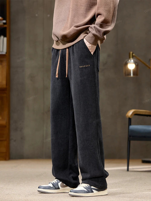 Person wearing dark blue velvet pants with a subtle brand logo, standing in a room with a gray wall and wooden furniture.