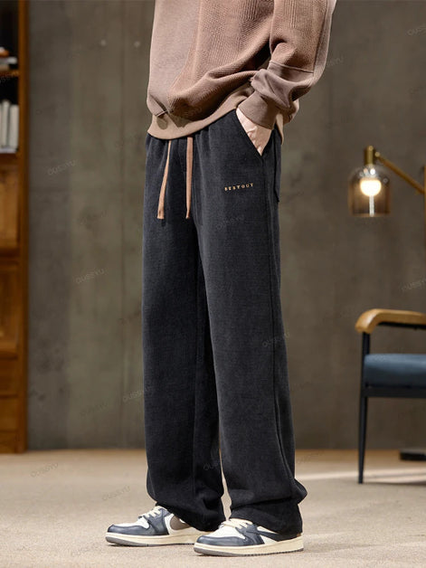 Person wearing dark blue velvet pants with a subtle brand logo, standing in a room with a gray wall and wooden furniture.