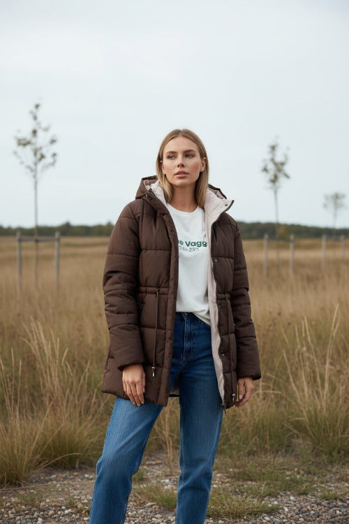 A woman wearing a dark brown puffer jacket with a light beige hood lining, styled with a white graphic t-shirt and blue jeans in a modern indoor setting.