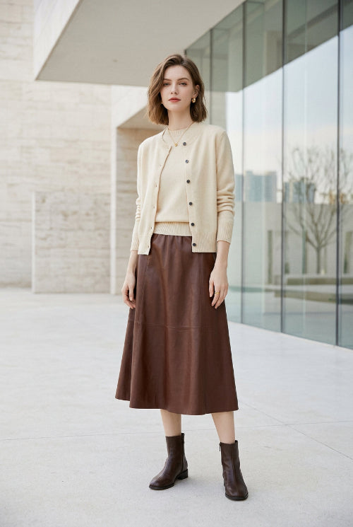 A person wearing a cream-colored knit cardigan over a matching cream sweater, paired with a brown faux-leather skirt, shown from the neck down in an indoor setting.