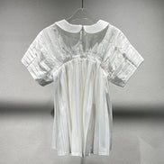 A white short-sleeve blouse with a Peter Pan collar, pleated bodice, and elasticized waist, hanging on a clear plastic hanger against a textured gray wall.