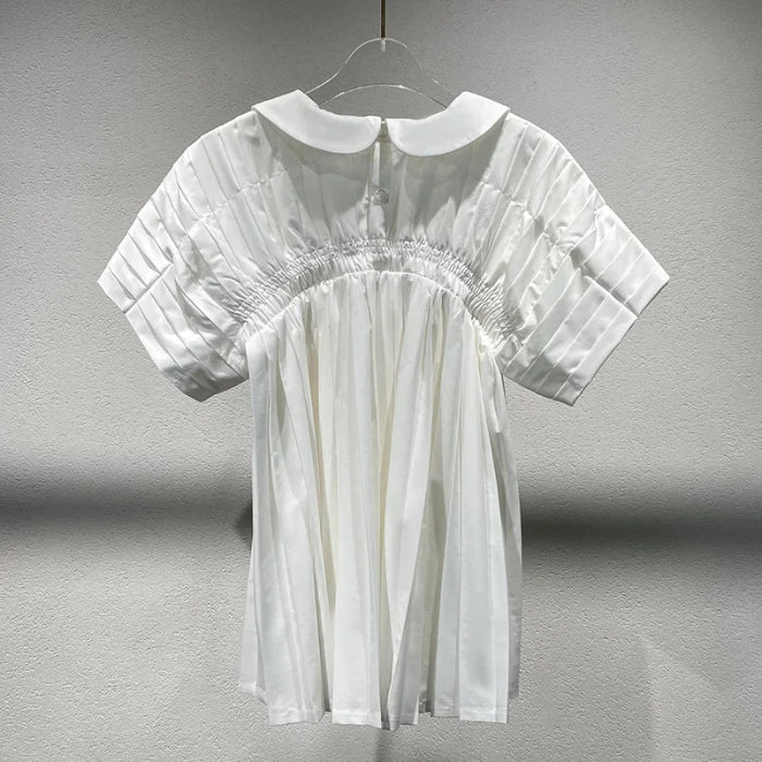 A white short-sleeve blouse with a Peter Pan collar, pleated bodice, and elasticized waist, hanging on a clear plastic hanger against a textured gray wall.