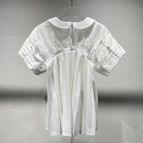 A white short-sleeve blouse with a Peter Pan collar, pleated bodice, and elasticized waist, hanging on a clear plastic hanger against a textured gray wall.