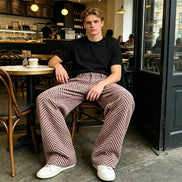 Man wearing brown and cream herringbone wide-leg pants, a black T-shirt, and white sneakers, seated inside a café