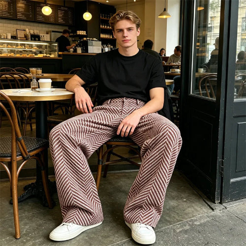 Man wearing brown and cream herringbone wide-leg pants, a black T-shirt, and white sneakers, seated inside a café