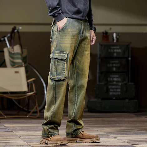 Side-angle view of a model wearing faded olive-brown denim cargo pants with a large flap thigh pocket and one hand in a side pocket, paired with brown boots.